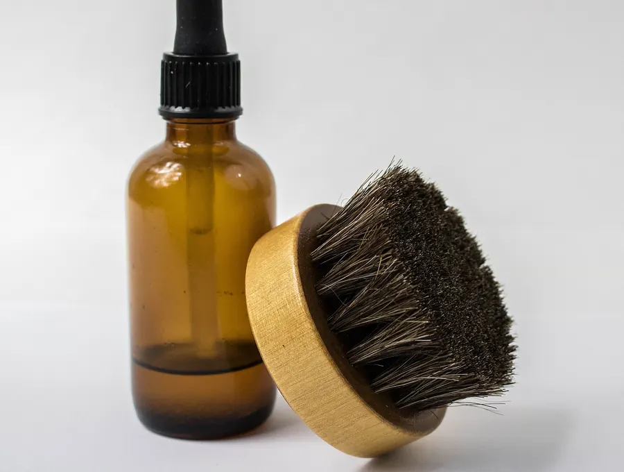 Amber dropper bottle of beard oil beside a round wooden beard brush.